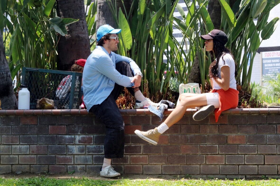 KJ Apa conversa com uma mulher desconhecida, ambas empoleiradas em um muro baixo de tijolos do lado de fora