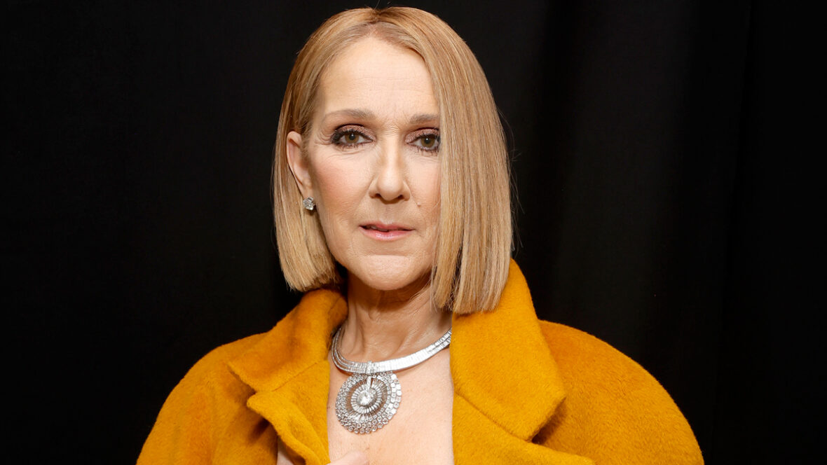 Celine Dion em close-up médio, vestindo um blazer laranja com um colar extravagante contra um fundo preto
