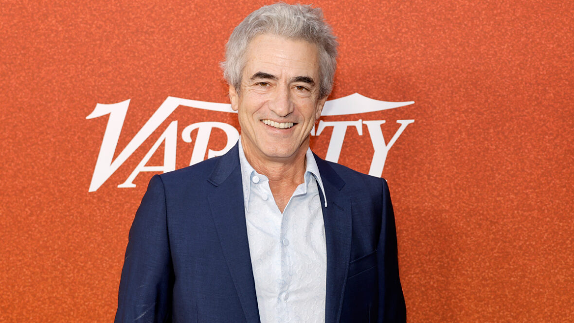 Dermot Mulroney sorri enquanto veste um terno em frente a um fundo laranja no evento Variety