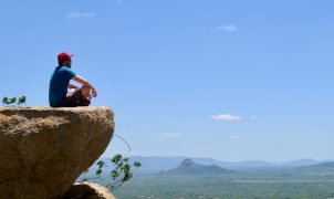man in blue t-shirt sitting on brown rock near body of water during daytime