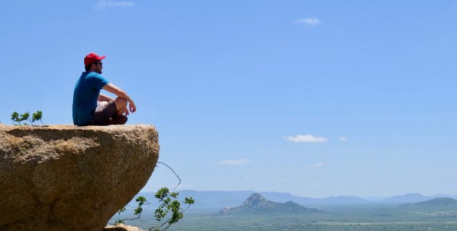 man in blue t-shirt sitting on brown rock near body of water during daytime