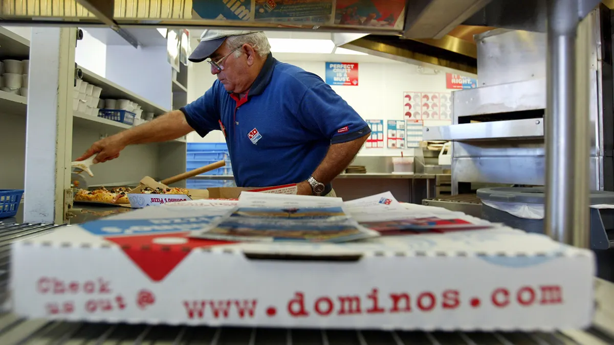 A Domino's employee cuts pizza slices in a box. In the foreground is a waiting box of pizza.