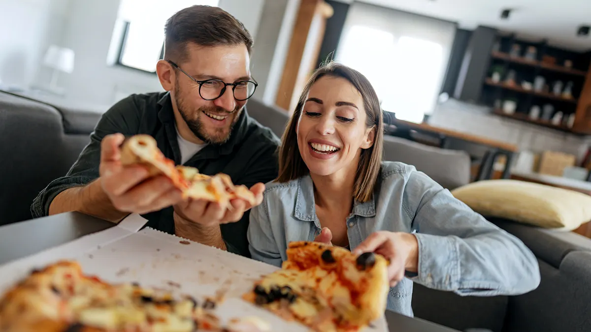 A man and a woman smile as they hold pizza from a delivery box at a home.