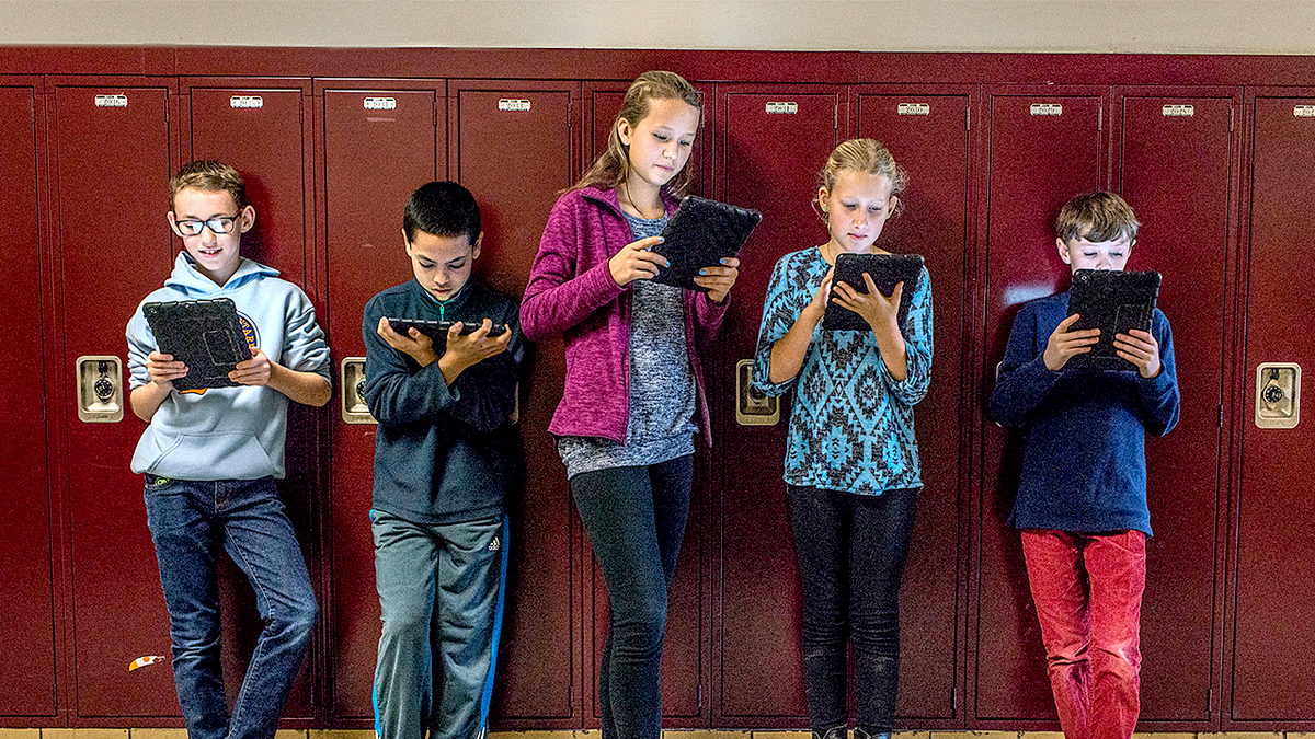 Students with tablets stand in front of red school lockers