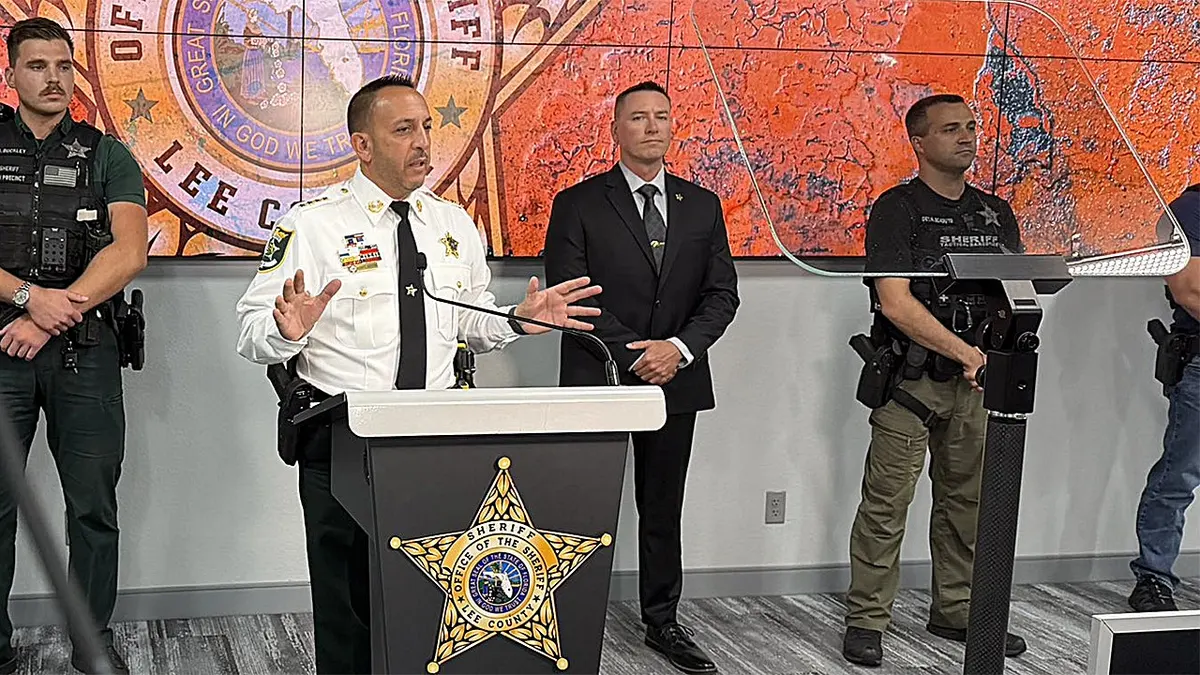 Lee County Sheriff Carmine Marceno speaks at a podium during a press conference while deputies and investigators stand behind him.