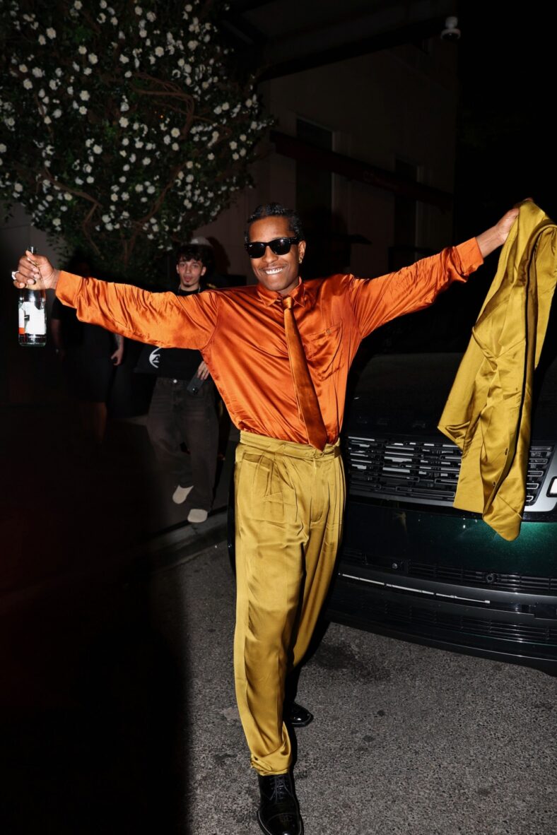 A$AP Rocky comemora a vitória dos Knicks nas ruas de Nova York enquanto veste uma camisa de cetim laranja brilhante e calças pregueadas, ele mantém os braços estendidos com uma garrafa na mão direita