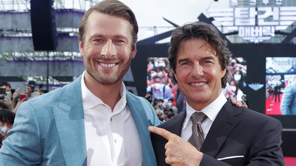 Glen Powell wearing white button down and blue teal blazer standing next to Tom Cruise pointing in black suit on red carpet