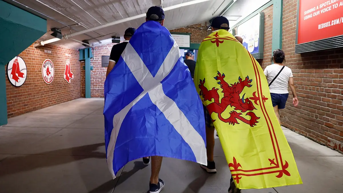 Two Scottish soccer fans walk through Fenway Park with flags draped over their backs.