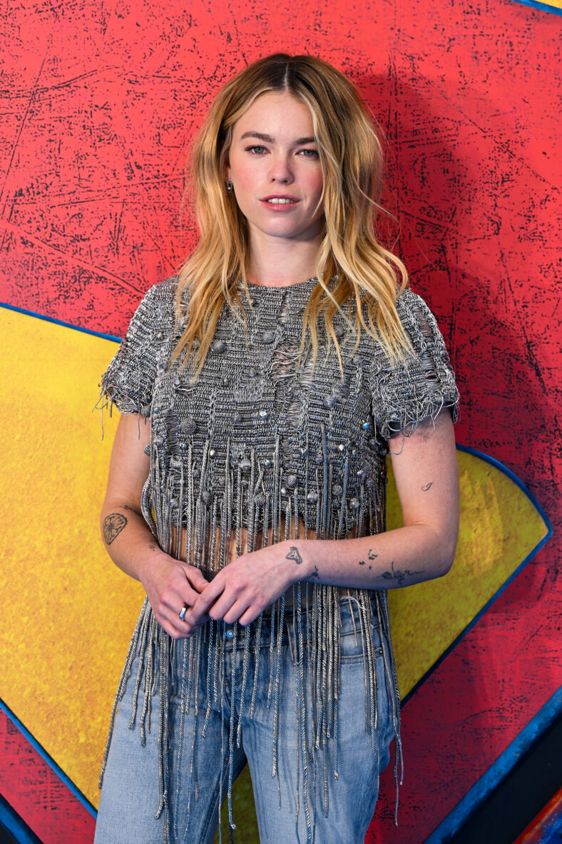 Milly Alcock posa contra um cenário de Supergirl vermelho, amarelo e azul enquanto veste um top texturizado com franjas cinza prateado, jeans e mostra várias tatuagens visíveis no braço.