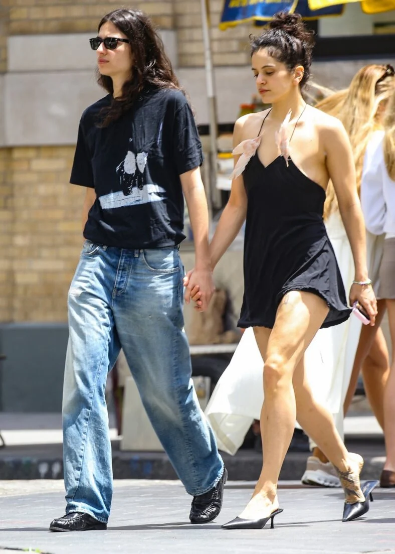 Rosália e Loli Bahia andam juntas de mãos dadas. Rosália usa um minivestido preto com o cabelo preso em um coque. Loli usa uma camiseta preta com óculos escuros.
