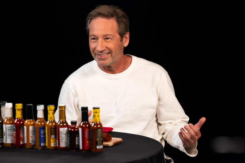 David Duchovny aparece no Hot Ones, sentado atrás de uma fila de molhos picantes enquanto veste um suéter branco.