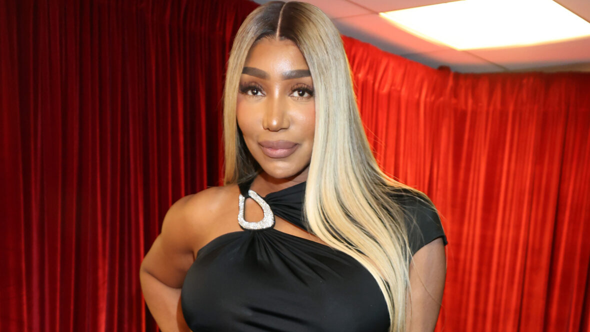 vazamentos de nene NeNe Leakes sorri enquanto posa diante de um cenário colorido em um evento. Ela usa longos cabelos loiros e um vestido preto com detalhes em renda transparente.