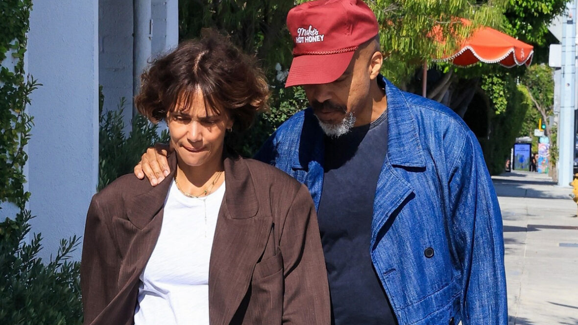 halle-berry-de-caça Halle Berry e o noivo Van Hunt andam de braços dados na calçada, com Van vestindo uma jaqueta azul brilhante e boné vermelho, enquanto Halle usa um blazer marrom grande e jeans