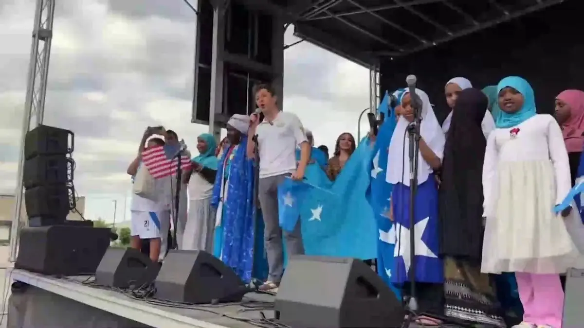 Minneapolis Mayor Jacob Frey speaks into a microphone on an outdoor stage surrounded by members of the Somali community holding blue Somali flags during a Somali Independence Day celebration.
