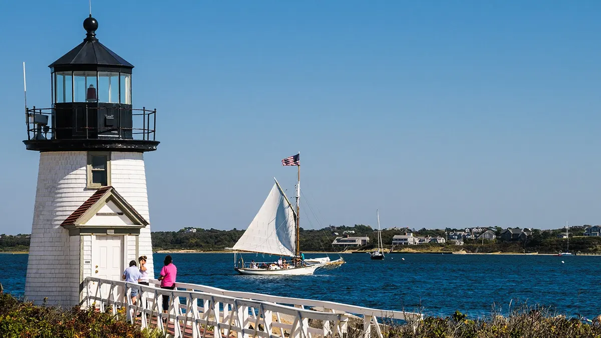 White lighthouse overlooking a blue harbor as a sailboat with a raised white sail passes by on a clear, sunny day.