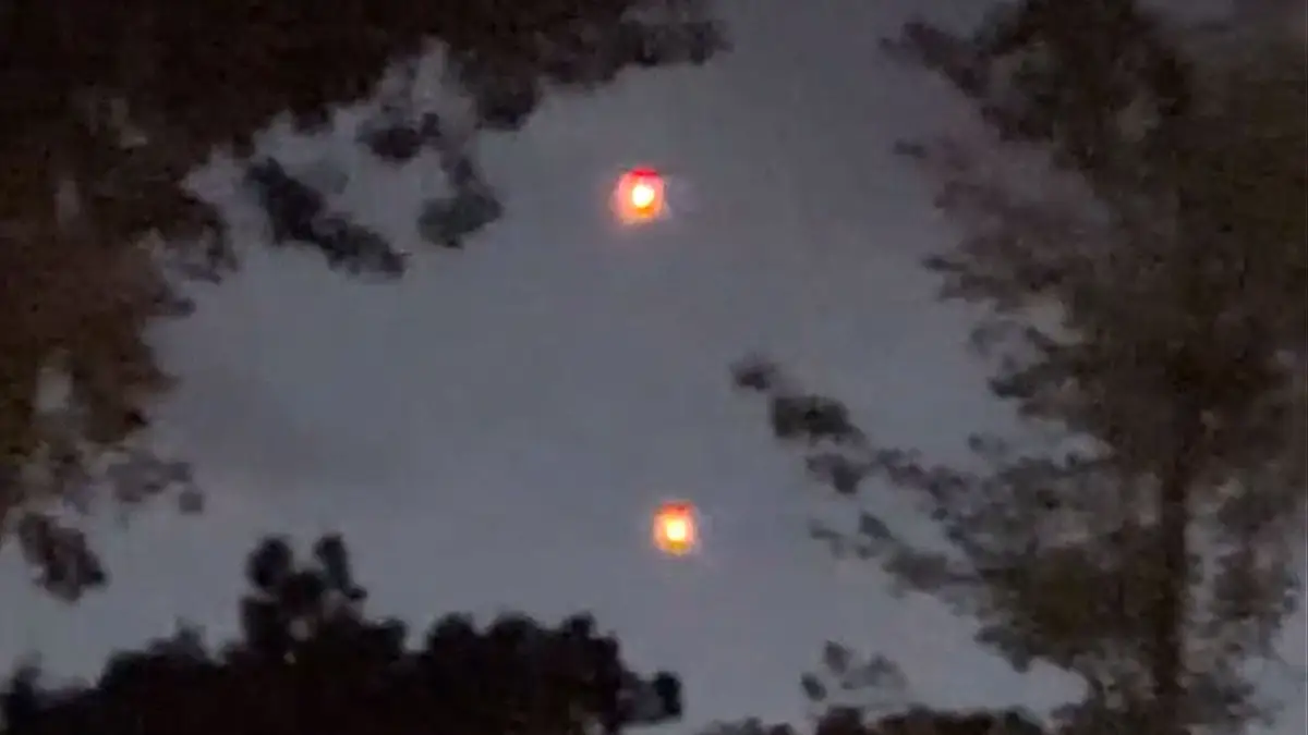 Two bright orbs hovering in tandem above a dark tree line in the night sky.