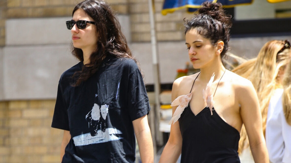 rosalia-mudança Rosália e Loli Bahia andam juntas de mãos dadas. Rosália usa um minivestido preto com o cabelo preso em um coque. Loli usa uma camiseta preta com óculos escuros.
