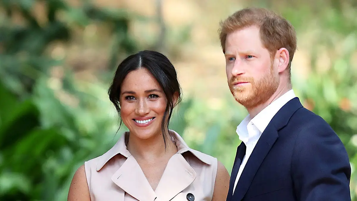A close-up of a smiling Meghan Markle and a stern-looking Prince Harry against a forest backdrop.