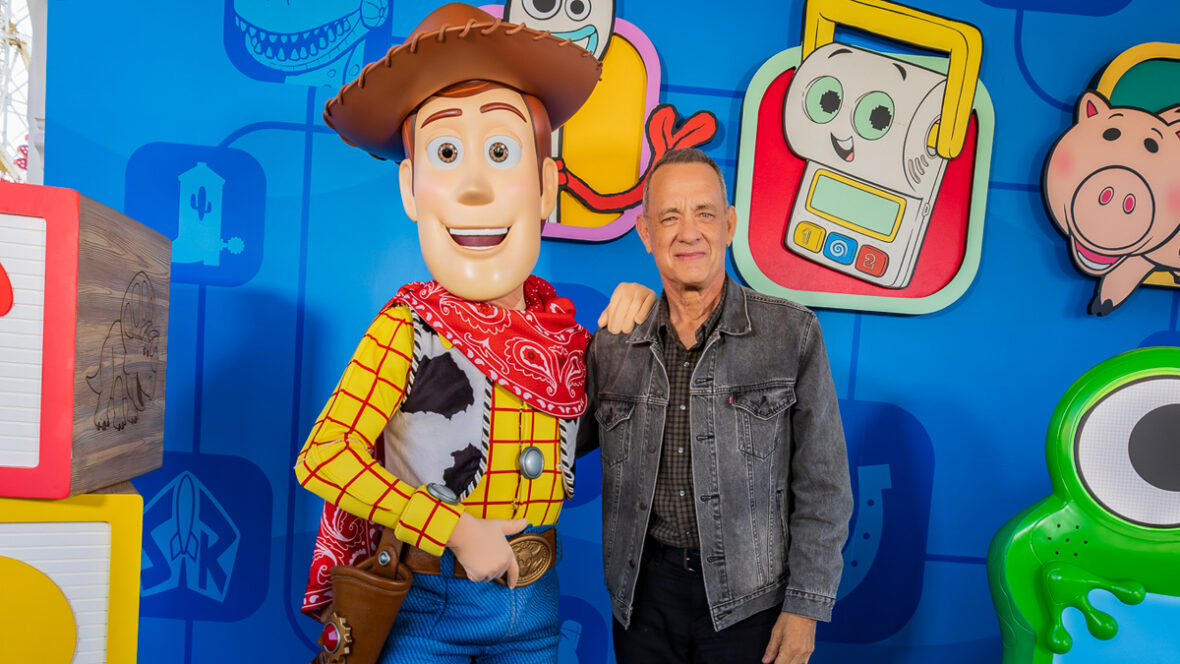 toy-story-6-tom-hanks Tom Hanks posa ao lado de uma grande figura de Woody em um evento Toy-Story com blocos de brinquedo coloridos, ilustrações de personagens e um fundo azul brilhante atrás deles.