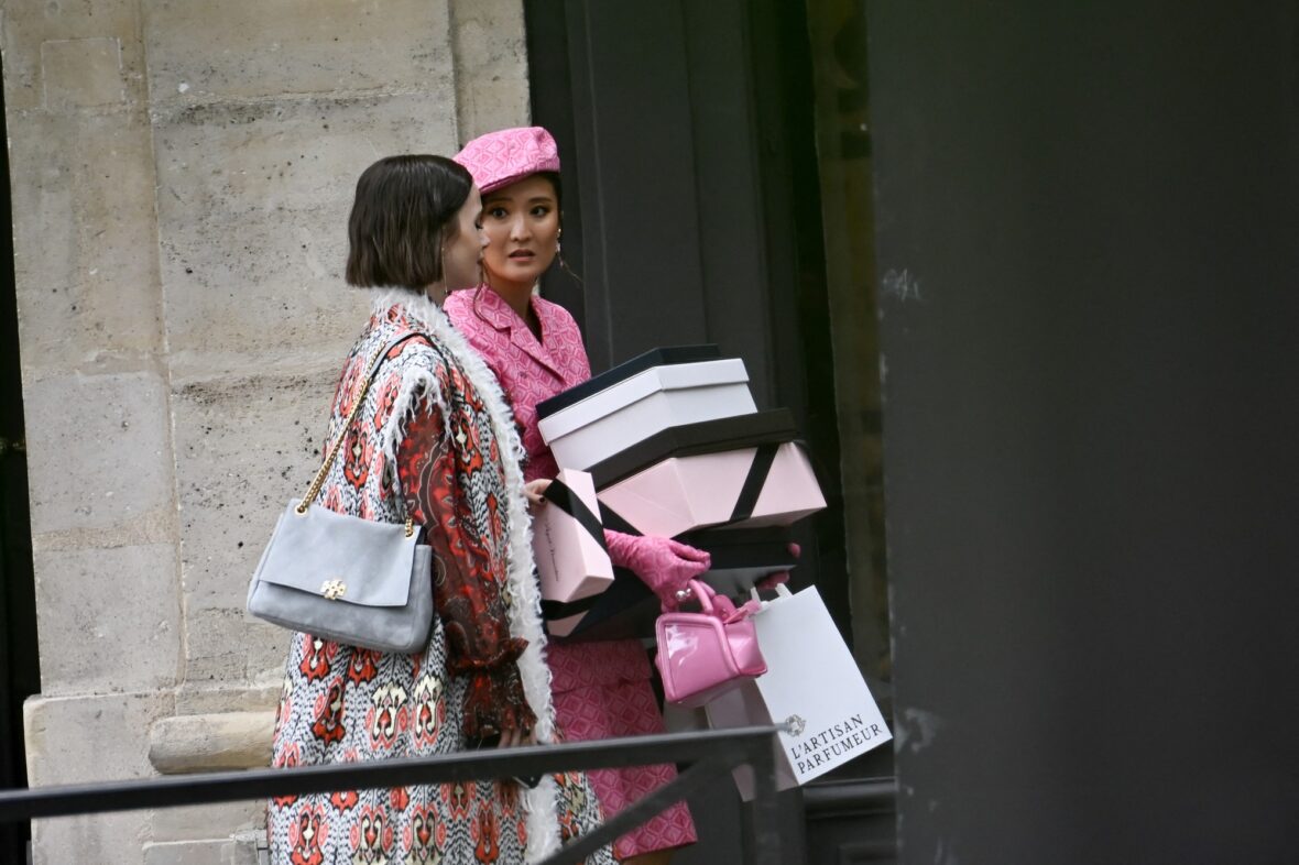 Lily Collins e Ashley Park filmam Emily em Paris em Paris, com Ashley vestindo uma roupa rosa brilhante e carregando caixas de compras enquanto Lily usa um casaco estampado.