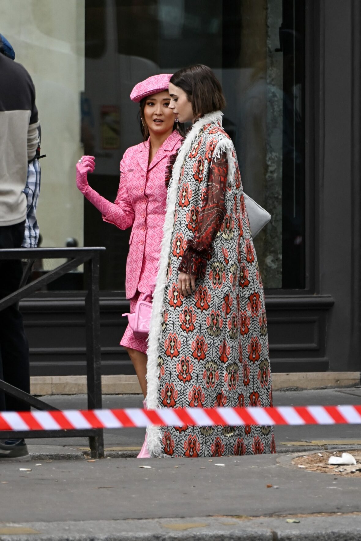 Ashley Park conversa com Lily Collins no set de Emily em Paris, com Ashley em uma boina rosa e terno combinando e Lily em um longo casaco estampado.