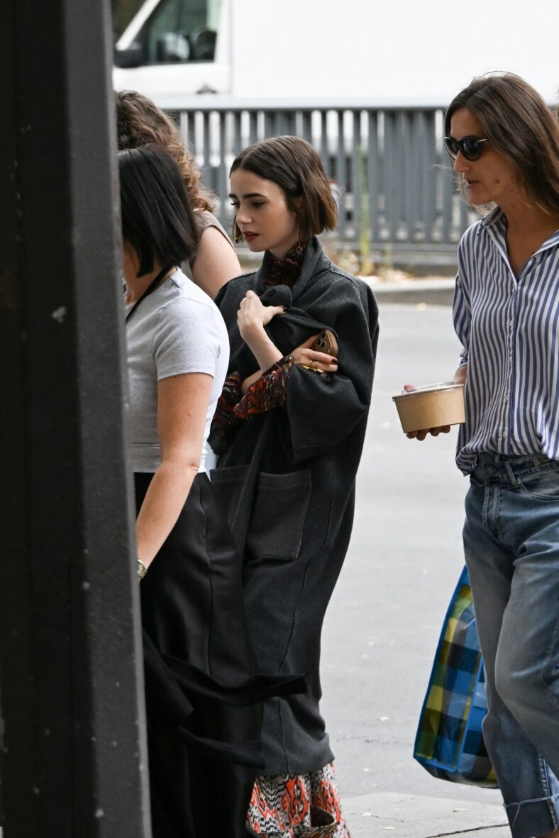Lily Collins caminha no set de Emily em Paris vestindo um casaco cinza sobre sua roupa estampada enquanto segura o telefone.
