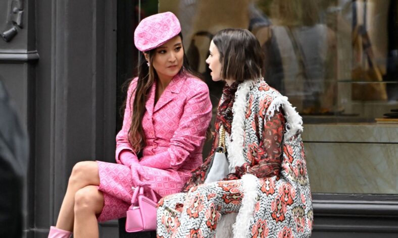 Lily Collins e Ashley Park sentam-se juntas enquanto filmam Emily em Paris, com Ashley em um look totalmente rosa e Lily vestindo um ousado casaco vermelho, preto e branco.