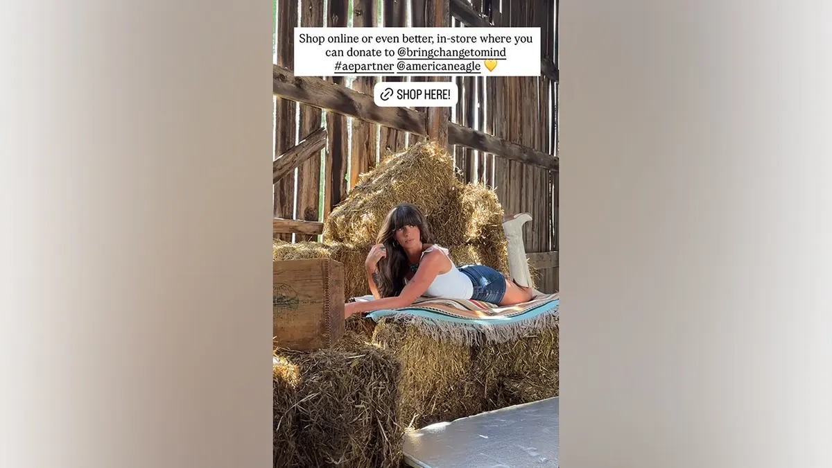 Ella Langley lies across stacked hay bales while wearing a white tank top, dark-wash denim shorts and white cowboy boots during an American Eagle photoshoot.