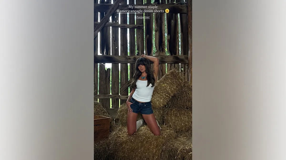 Ella Langley kneels atop stacked hay bales while wearing a white tank top, dark-wash denim shorts and white cowboy boots during an American Eagle photoshoot.