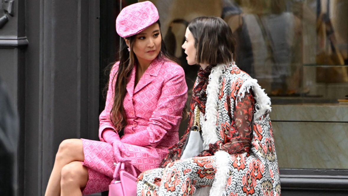 Lily Collins e Ashley Park sentam-se juntas enquanto filmam Emily em Paris, com Ashley em um look totalmente rosa e Lily vestindo um ousado casaco vermelho, preto e branco.