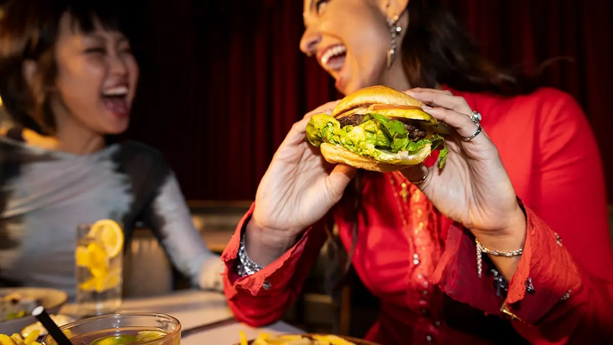 A woman holds a burger and laughs with a friend.