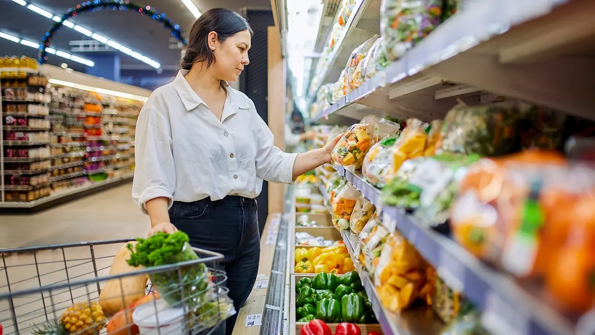 Woman shops for fresh vegetables in a supermarket produce aisle with a shopping cart.