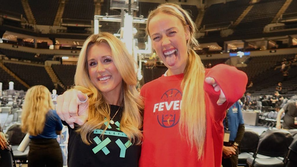 Riley Gaines and Indiana Fever guard Sophie Cunningham posing together on the basketball court, smiling and pointing at the camera inside a packed arena.