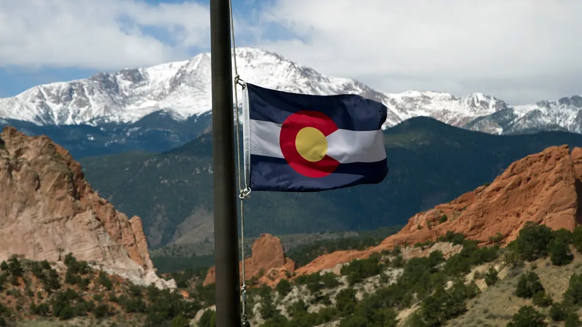 Colorado state flag waves with Pikes Peak and Garden of the Gods visible in the background.