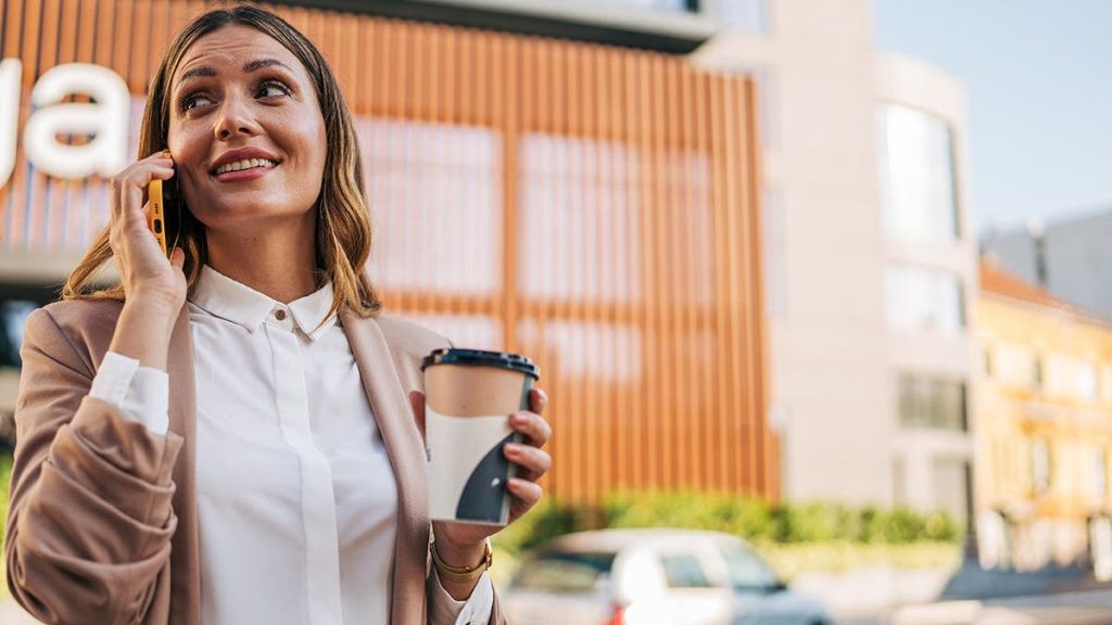 Businesswoman smiles while talking on phone and holding coffee cup outside office building.