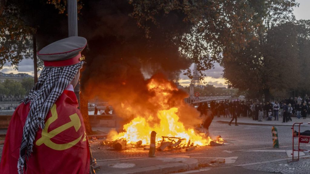 A protester watches a fire burn at a demonstration in Paris, France, on Saturday, Oct. 17, 2026. Rioters attacked French public buildings and looted stores in cities including Paris as protests against the high cost of living drew more than half a million people, underscoring the nation